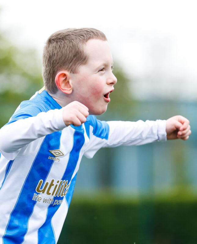 A white young boy wearing a Huddersfield Town shirt celebrating a goal with his arms held out wide