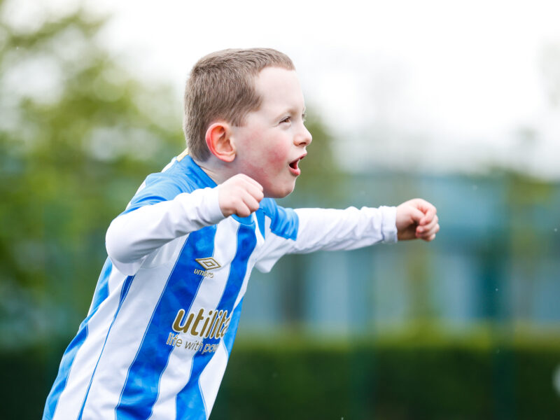 A white young boy wearing a Huddersfield Town shirt celebrating a goal with his arms held out wide