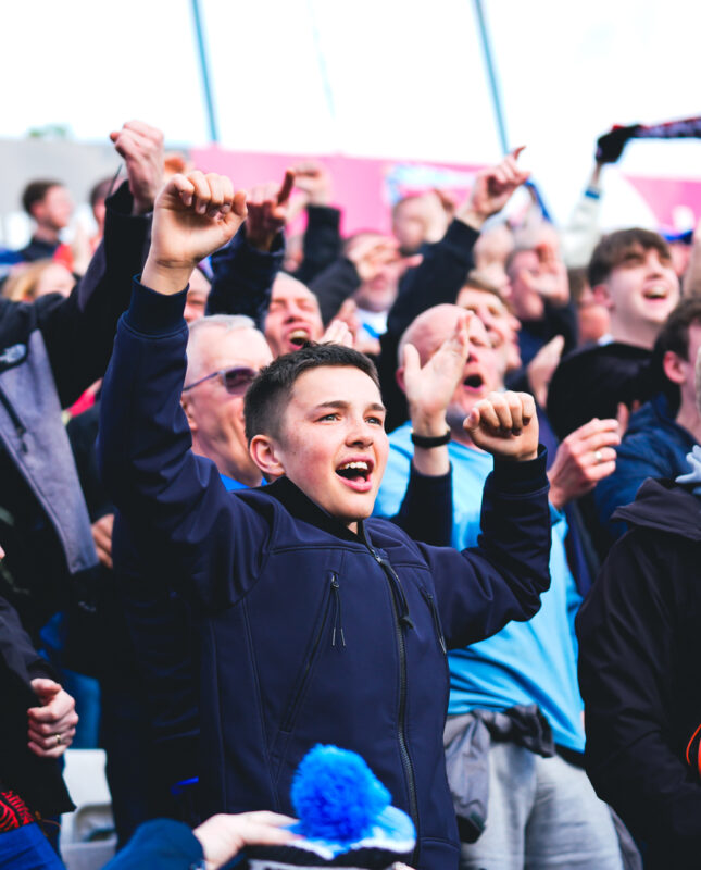 Male Huddersfield Town fans celebrating with their hands in the air in the stands as Huddersfield score away to Stockport County